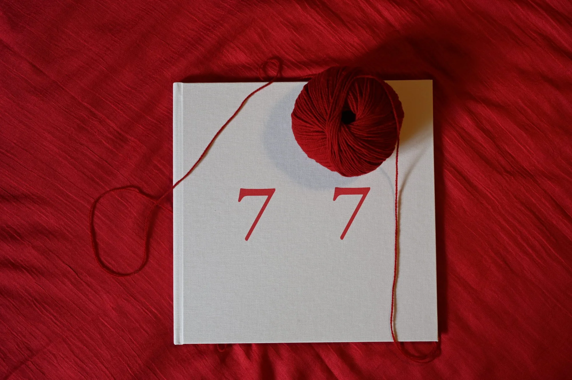 7 7 — the pale linen cover, the title in red, a ball of red wool with its thread wandering across the cloth.